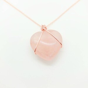 May include: A rose quartz heart pendant necklace. The heart-shaped stone is a soft pink color and is wrapped in copper-colored wire. The necklace chain is also copper-colored and delicate. The background is white.