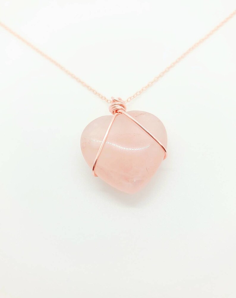 May include: A rose quartz heart pendant necklace. The heart-shaped stone is a soft pink color and is wrapped in copper-colored wire. The necklace chain is also copper-colored and delicate. The background is white.