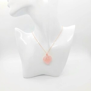May include: A rose gold necklace with a heart-shaped pendant. The pendant is a light pink stone wrapped in rose gold wire. The necklace is displayed on a white mannequin bust.