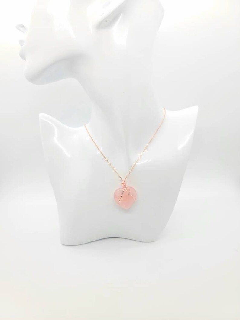 May include: A rose gold necklace with a heart-shaped pendant. The pendant is a light pink stone wrapped in rose gold wire. The necklace is displayed on a white mannequin bust.