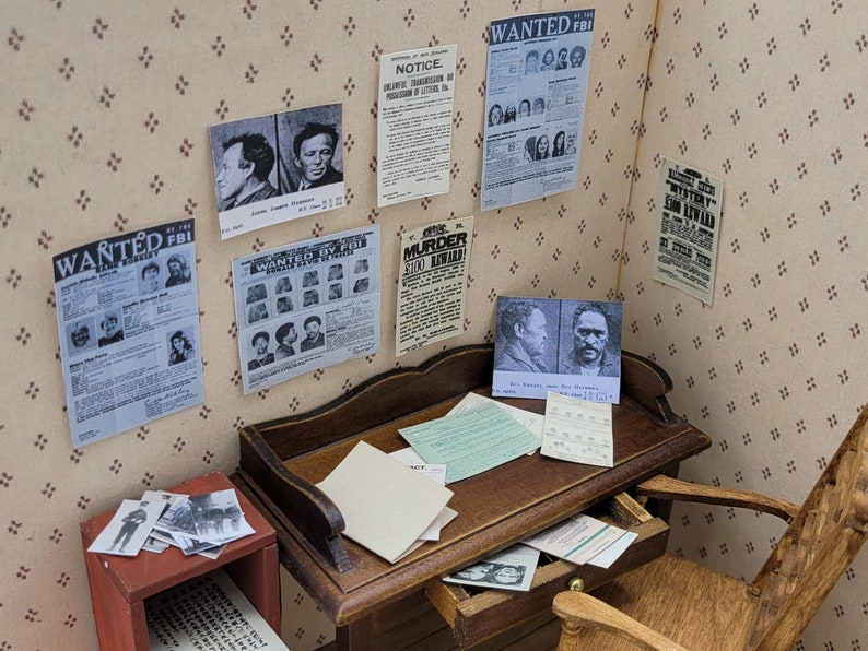May include: A miniature detective's office with a wooden desk, a chair, and a small red shelf. The walls are covered in wanted posters with black and white photos of people. The posters have the text "WANTED BY THE FBI" and "MURDER - $100 REWARD!"
