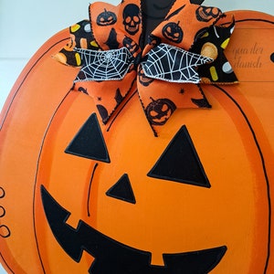 Op de afbeelding: Oranje en zwarte Halloween-pompoen deurhanger met een zwart-witte strik met een spinnenwebpatroon. De pompoen heeft een zwart geschilderd gezicht met een brede glimlach en driehoekige ogen.