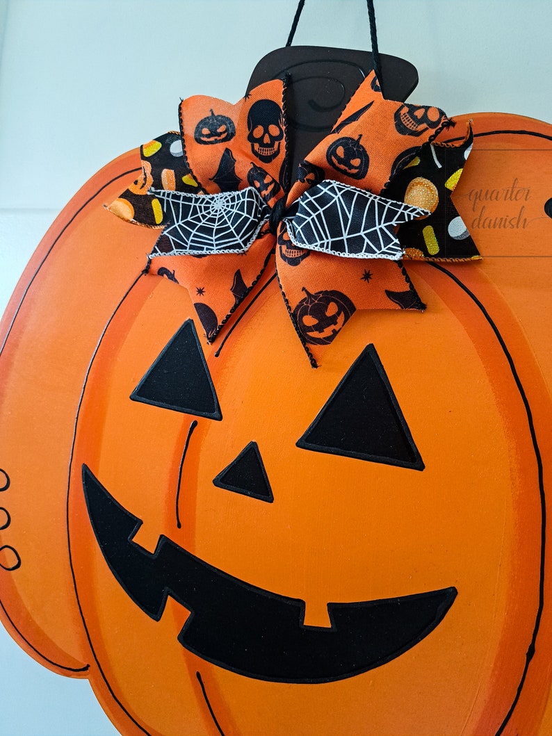 Op de afbeelding: Oranje en zwarte Halloween-pompoen deurhanger met een zwart-witte strik met een spinnenwebpatroon. De pompoen heeft een zwart geschilderd gezicht met een brede glimlach en driehoekige ogen.