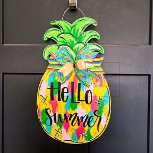May include: A yellow pineapple-shaped door hanger with a green top and a colorful, abstract design. The text "Hello Summer" is written in black on the front of the hanger.