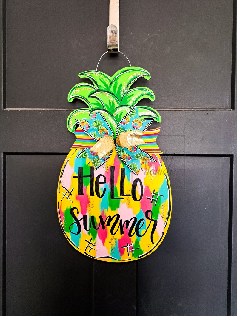 May include: A yellow pineapple-shaped door hanger with a green top and a colorful, abstract design. The text "Hello Summer" is written in black on the front of the hanger.