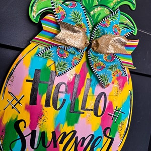 May include: A yellow pineapple-shaped wooden door hanger with a colorful abstract paint design and the text "Hello Summer" in black. The pineapple has a green top with gold glitter and a blue and white patterned bow with gold ribbon.