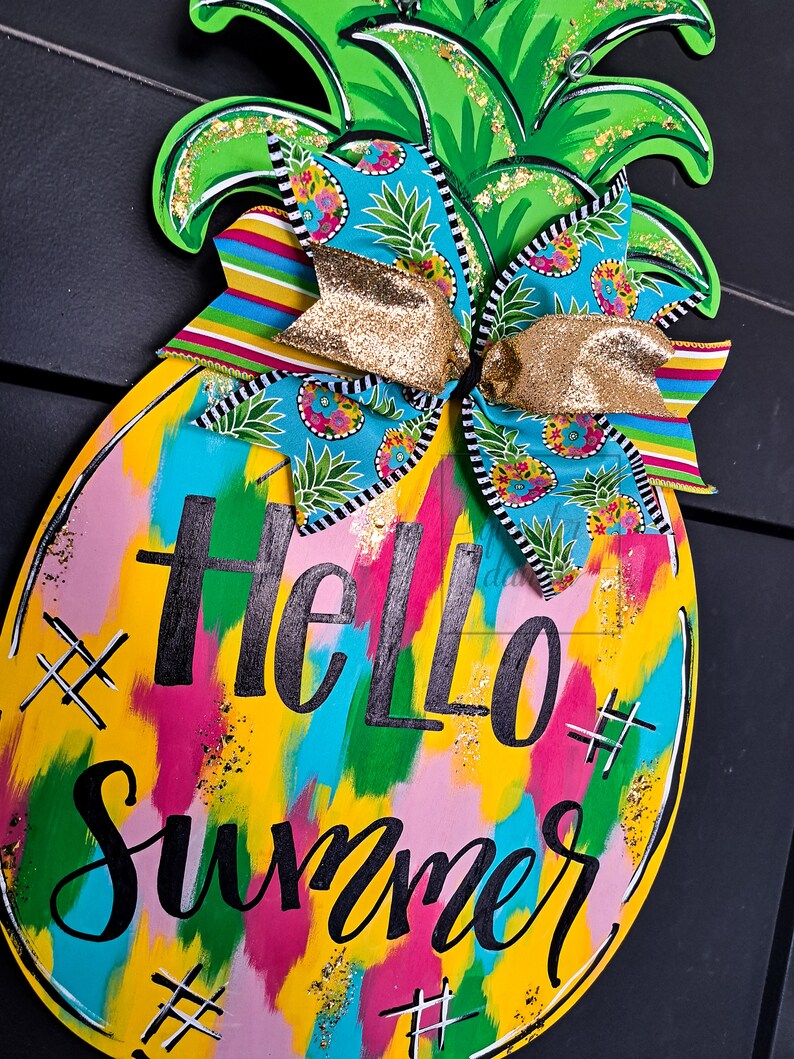 May include: A yellow pineapple-shaped wooden door hanger with a colorful abstract paint design and the text "Hello Summer" in black. The pineapple has a green top with gold glitter and a blue and white patterned bow with gold ribbon.