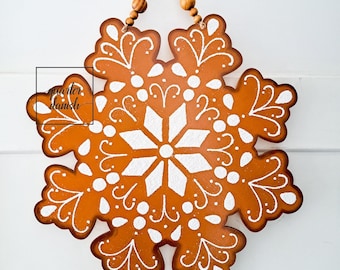 May include: A brown wooden snowflake ornament with a white painted design. The snowflake has a loop at the top for hanging.