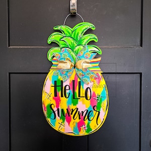 May include: A yellow pineapple-shaped door hanger with a green top and a colorful bow. The pineapple has the text "Hello Summer" written on it in black.