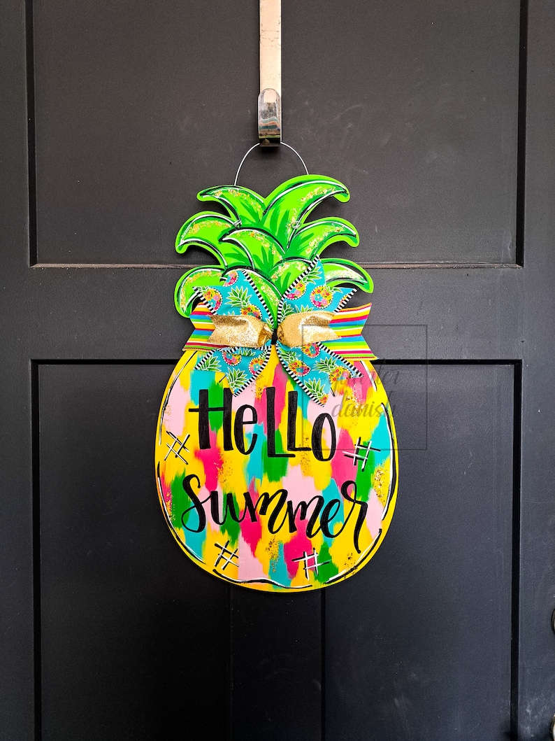 May include: A yellow pineapple-shaped door hanger with a green top and a colorful bow. The pineapple has the text "Hello Summer" written on it in black.