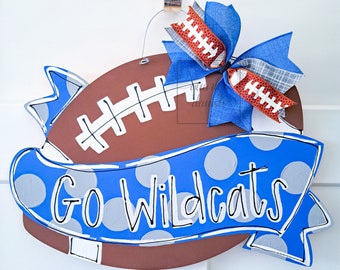 May include: A brown football-shaped door hanger with a blue and white polka dot banner that says "Go Wildcats". A blue and white bow with a brown football ribbon is attached to the top.