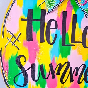 May include: A round wooden sign with a yellow background and colorful abstract brushstrokes. The sign says "Hello Summer" in black script lettering with gold glitter accents.