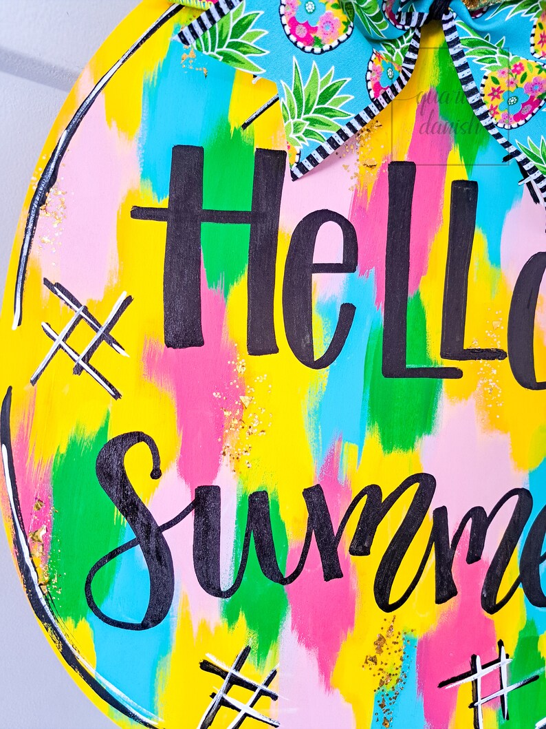 May include: A round wooden sign with a yellow background and colorful abstract brushstrokes. The sign says "Hello Summer" in black script lettering with gold glitter accents.