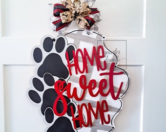 May include: A black and white checkered door hanger with a red "Home Sweet Home" inscription. The hanger is decorated with black paw prints and a red and black bow.