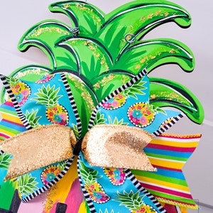 May include: A wooden pineapple door hanger with a bright green pineapple and a colorful bow. The bow is made of blue and white fabric with a pineapple pattern and a gold glitter ribbon. The pineapple has gold glitter accents. The door hanger says "hello" in colorful letters.