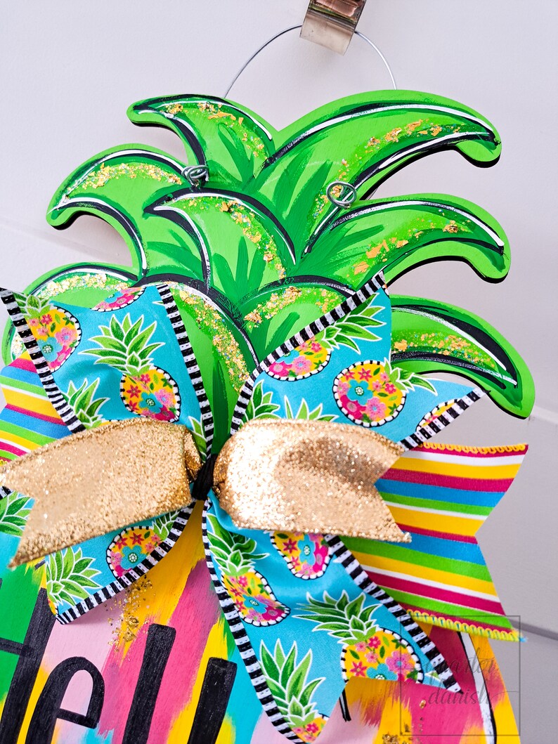 May include: A wooden pineapple door hanger with a bright green pineapple and a colorful bow. The bow is made of blue and white fabric with a pineapple pattern and a gold glitter ribbon. The pineapple has gold glitter accents. The door hanger says "hello" in colorful letters.