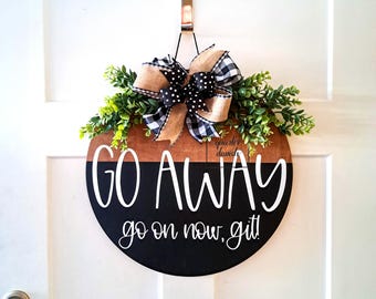 May include: A black and brown round wooden door hanger with the text "GO AWAY go on now, git!" in white lettering. The hanger is decorated with a black and white checkered ribbon and green leaves.