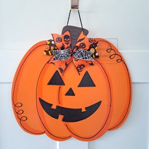 Op de afbeelding: Oranje en zwarte Halloween-deurhanger in de vorm van een pompoen met een zwart pompoengezicht en een zwart-witte strik met een spinnenwebontwerp.