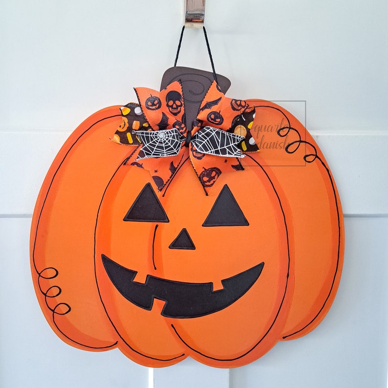 Op de afbeelding: Oranje en zwarte Halloween-deurhanger in de vorm van een pompoen met een zwart pompoengezicht en een zwart-witte strik met een spinnenwebontwerp.