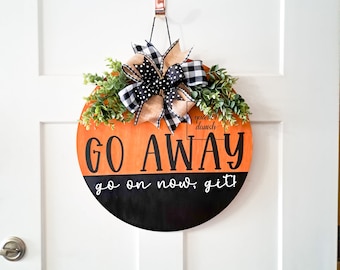May include: A round wooden door hanger with a black and orange pumpkin design. The top half of the pumpkin is orange and says "GO AWAY" in black lettering. The bottom half of the pumpkin is black and says "go on now, git!" in white lettering. The hanger is decorated with a black and white checkered bow and green leaves.