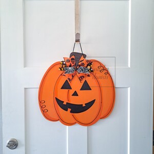Op de afbeelding: Een houten oranje pompoen deurhanger met een zwart jack-o'-lantern gezicht en een zwart-witte strik met een spinnenweb ontwerp.