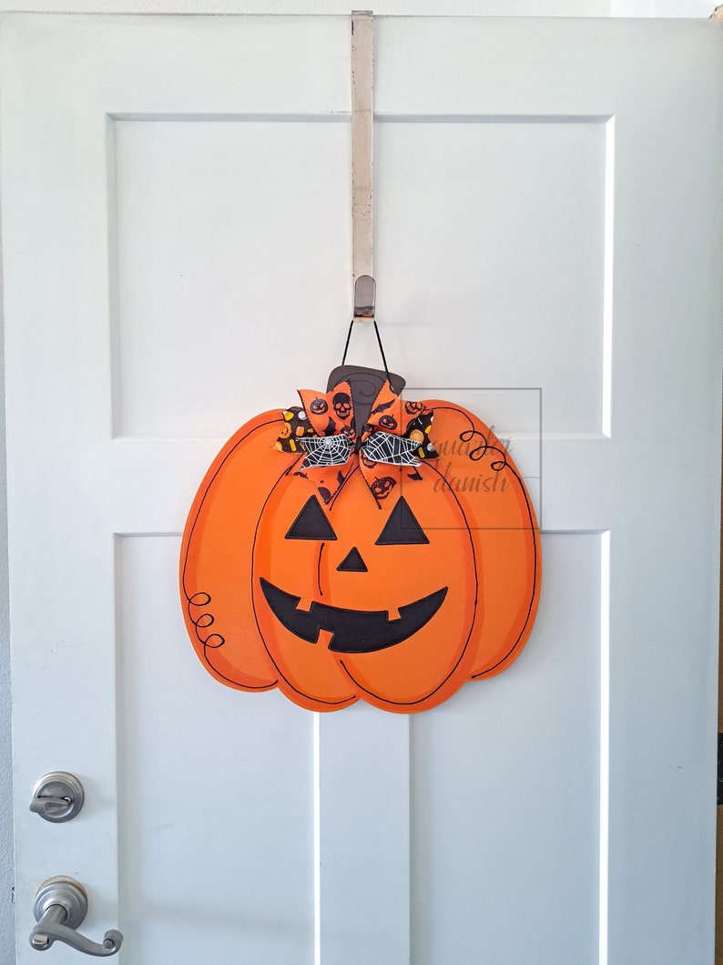 Op de afbeelding: Een houten oranje pompoen deurhanger met een zwart jack-o'-lantern gezicht en een zwart-witte strik met een spinnenweb ontwerp.