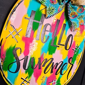 May include: A round wooden sign with a yellow background and colorful abstract brushstrokes. The sign says "Hello Summer" in black script with gold glitter accents. The sign is decorated with a blue and white pineapple patterned bow.