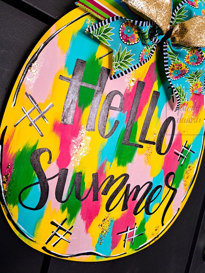 May include: A round wooden sign with a yellow background and colorful abstract brushstrokes. The sign says "Hello Summer" in black script with gold glitter accents. The sign is decorated with a blue and white pineapple patterned bow.