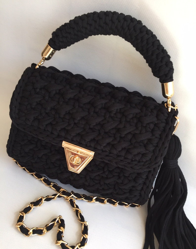 May include: A black crocheted handbag with a gold chain strap and a gold clasp. The bag has a black tassel hanging from the side.