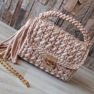 May include: A beige crocheted handbag with a gold clasp and a long tassel. The bag has a chain strap and a crocheted top handle.