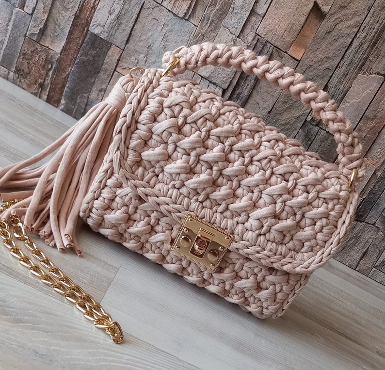 May include: A beige crocheted handbag with a gold clasp and a long tassel. The bag has a chain strap and a crocheted top handle.