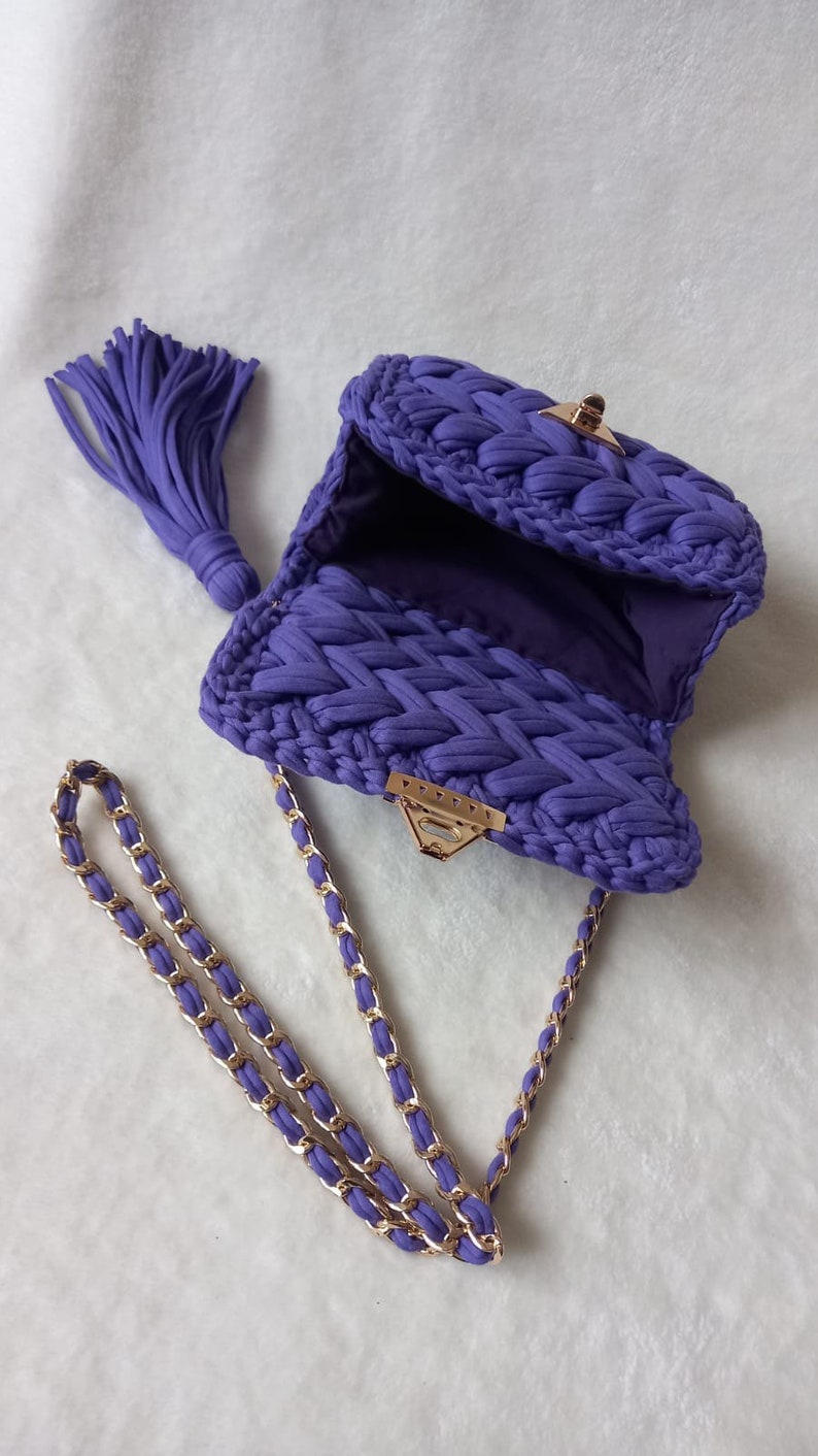 May include: A purple crocheted handbag with a gold chain strap and a tassel. The bag has a clasp closure.