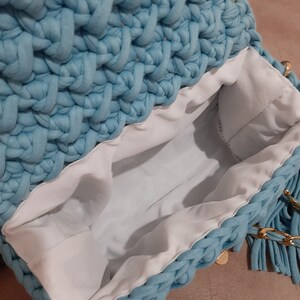 May include: A light blue crocheted bag with a white lining and a gold chain strap. The bag has a fringe detail on the bottom.