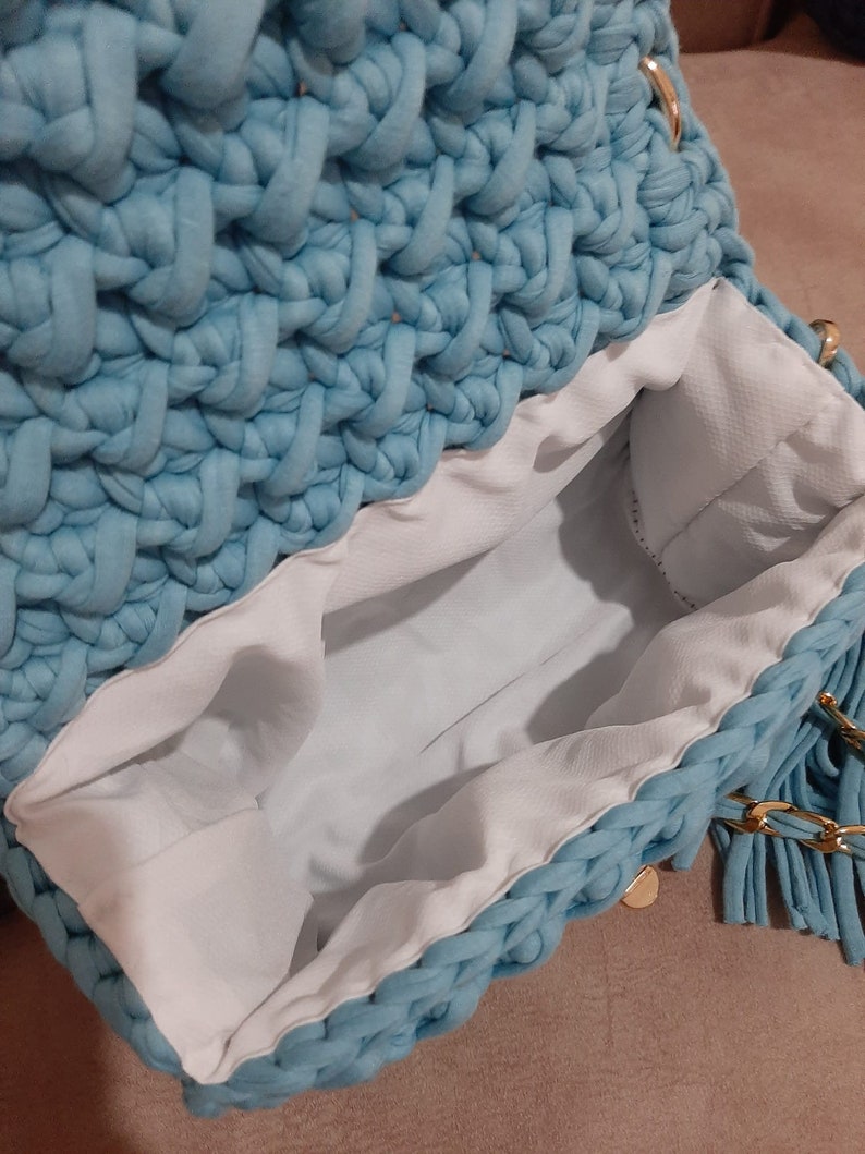 May include: A light blue crocheted bag with a white lining and a gold chain strap. The bag has a fringe detail on the bottom.