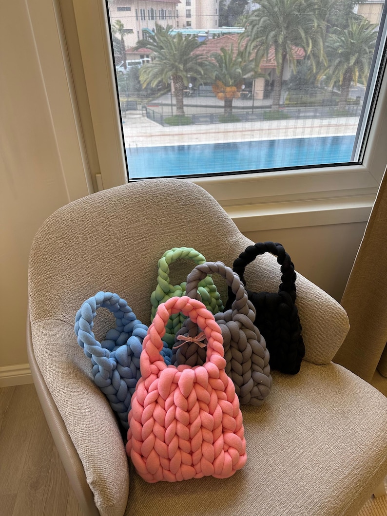 Puede incluir: Cinco bolsas de mano de punto en diferentes colores, incluyendo rosa, azul, gris, verde y negro. Las bolsas est&aacute;n sentadas en una silla frente a una ventana con vista a una piscina.
