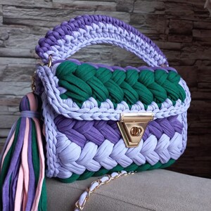 May include: A crocheted handbag with a green, purple, and light purple design. The bag has a gold clasp and a matching tassel.