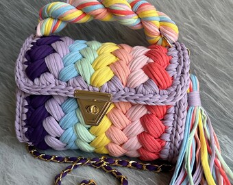 May include: A rainbow crocheted handbag with a braided handle and a gold clasp. The bag has a chain strap and a tassel made of the same crocheted yarn.