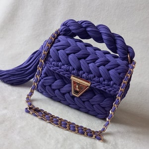 May include: A purple knitted handbag with a braided handle and a gold chain strap. The bag has a gold clasp and a tassel.