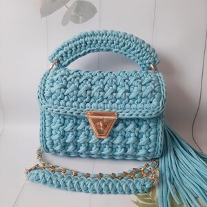 May include: A light blue crocheted handbag with a gold chain strap and a tassel. The bag has a flap closure with a gold clasp.