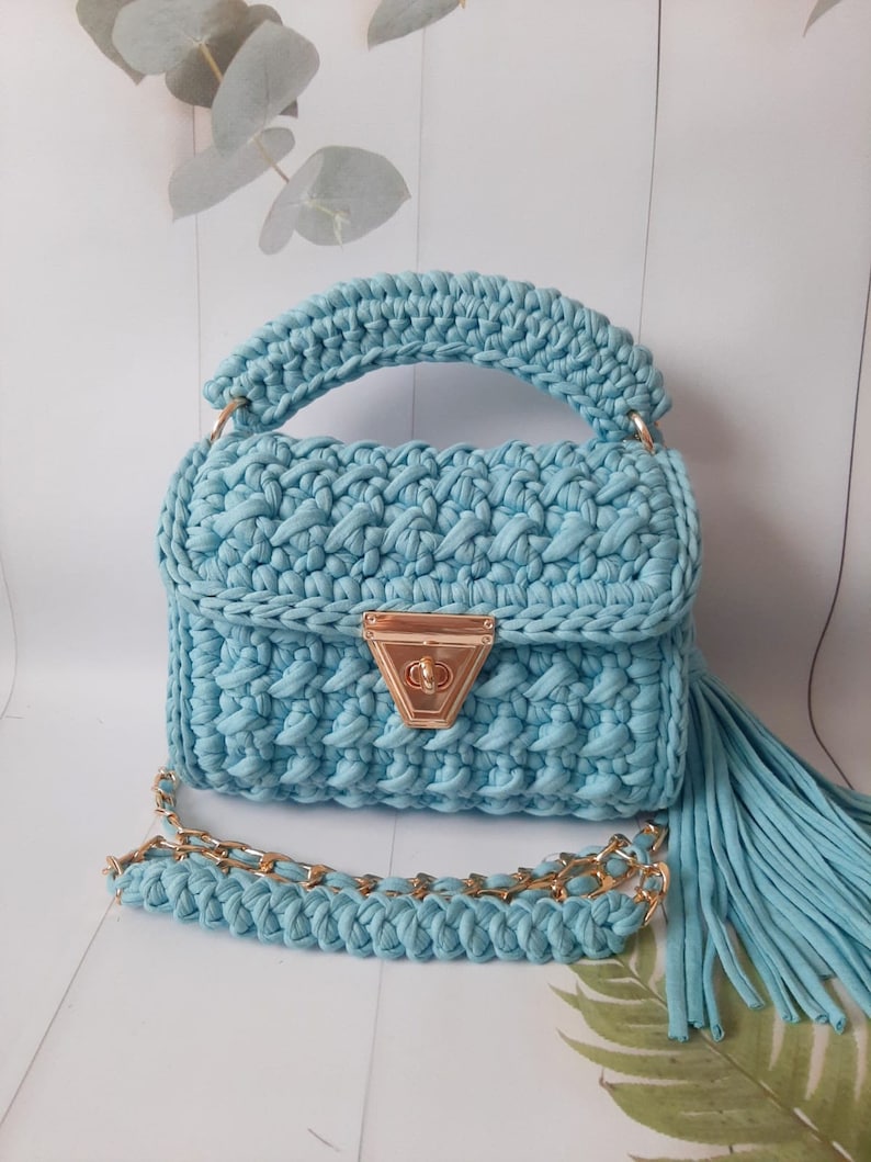 May include: A light blue crocheted handbag with a gold chain strap and a tassel. The bag has a flap closure with a gold clasp.