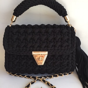 May include: A black crocheted handbag with a gold chain strap and a gold clasp. The bag has a tassel on the side.