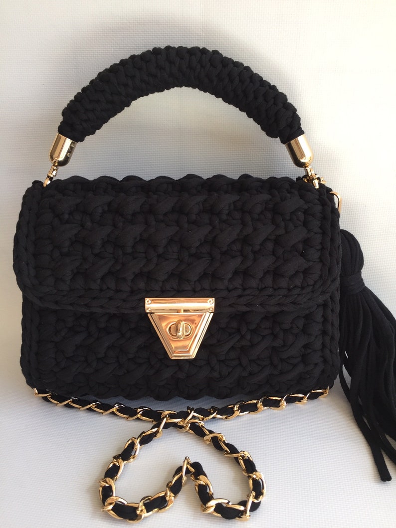 May include: A black crocheted handbag with a gold chain strap and a gold clasp. The bag has a tassel on the side.