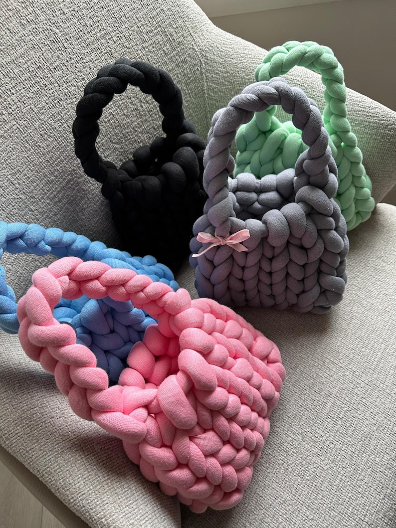 Puede incluir: Cuatro bolsas de mano de punto en diferentes colores: negro, azul claro, lavanda y rosa. Las bolsas est&aacute;n hechas de hilo grueso y tienen un dise&ntilde;o simple y minimalista.