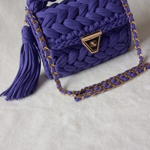 May include: A purple knitted handbag with a gold chain strap and a tassel. The bag has a braided handle and a gold clasp.