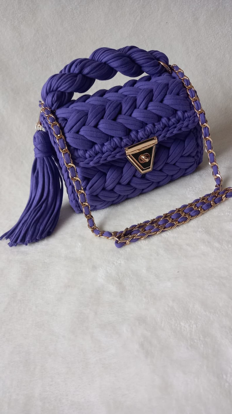 May include: A purple knitted handbag with a gold chain strap and a tassel. The bag has a braided handle and a gold clasp.