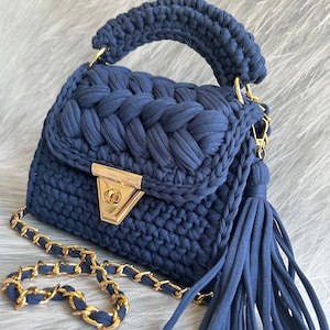 May include: A small, navy blue crocheted handbag with a gold chain strap and a tassel. The bag has a top handle and a gold clasp.