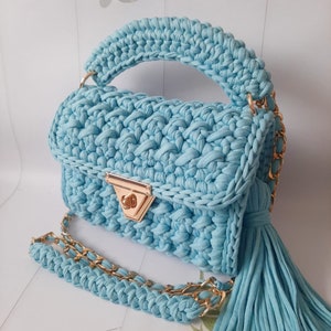May include: A light blue crocheted handbag with a gold chain strap and a tassel. The bag has a top handle and a flap closure with a gold clasp.