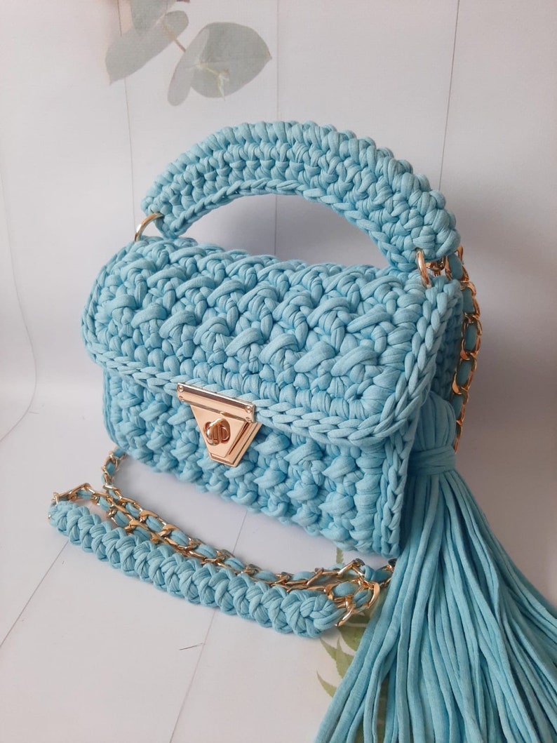 May include: A light blue crocheted handbag with a gold chain strap and a tassel. The bag has a top handle and a flap closure with a gold clasp.
