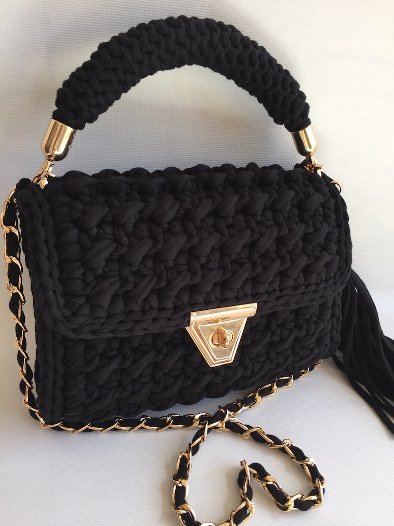 May include: A black crocheted handbag with a gold chain strap and a gold clasp. The bag has a top handle and a tassel.