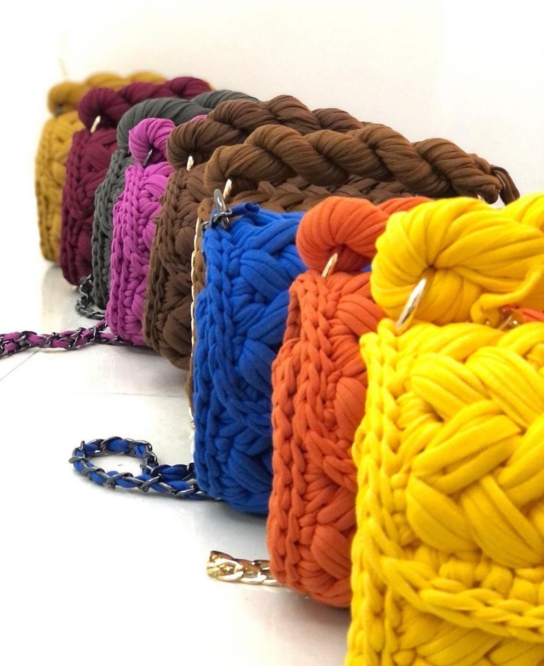 May include: A row of colourful crocheted handbags with chain straps. The bags are in shades of mustard yellow, burgundy, grey, pink, blue, orange, and yellow.